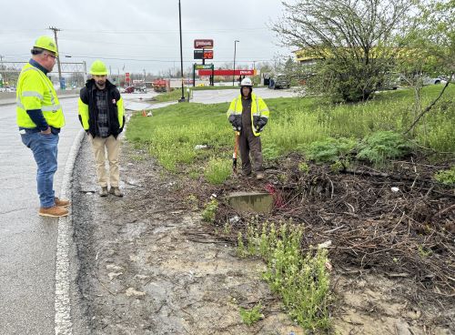 Three men in yellow, high-visibility jackets look at a ditch on the side of Frontage Road, which will soon be resurfaced and widened.